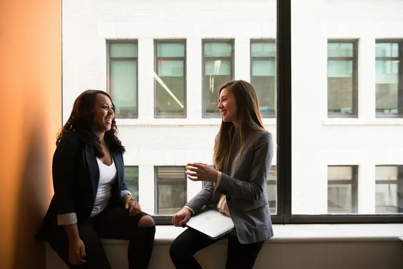 Zwei Frauen sprechen lachend auf einer Fensterbank im Büro