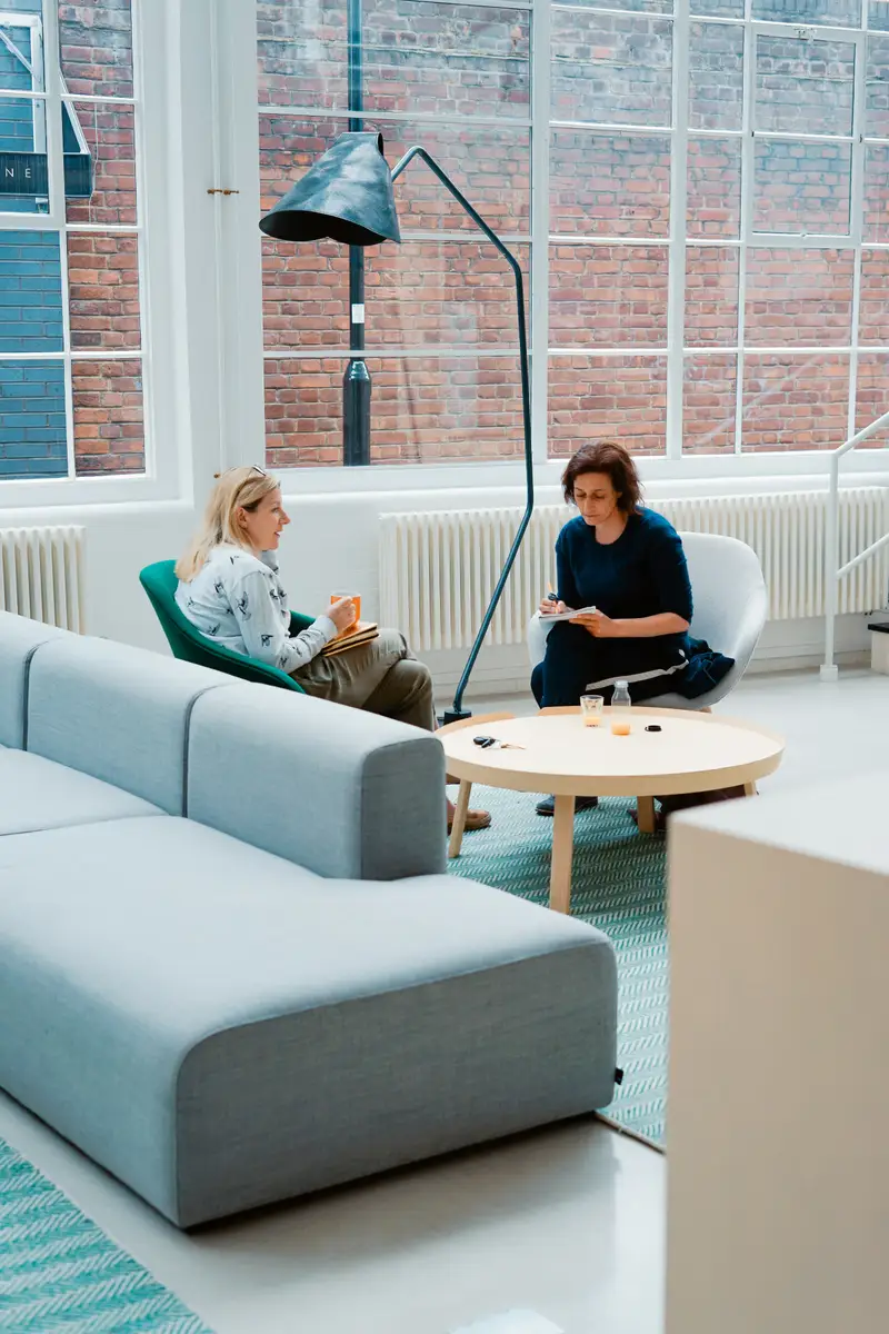 Zwei Frauen im Gespräch in einem hellen Raum mit Sofa und großen Fenstern