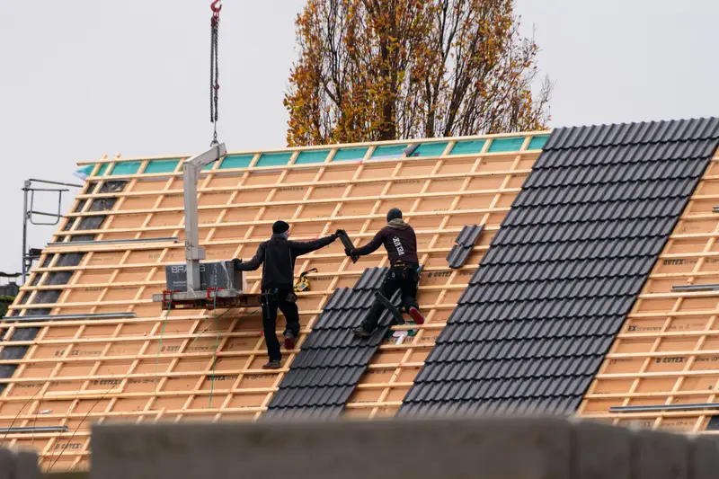 Zwei Dachdecker decken ein Steildach mit dunklen Ziegeln neu ein, im Hintergrund ein Herbstbaum