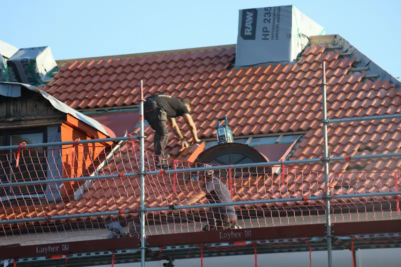 Dachdecker repariert ein rotes Ziegeldach, das Gerüst ist mit Fangnetz gesichert