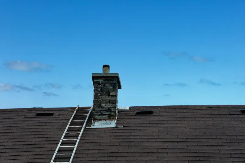 Kamin auf einem dunklen Dach, daran lehnt eine Dachleiter